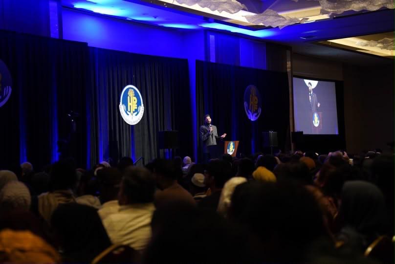 Speaker on stage with blue lighting, projection, and a full audience